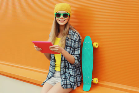 Happy modern stylish young woman using digital tablet computer, cool creative freelancer girl smiles in colorful trendy clothes with skateboard on city street on orange wall backgroundの写真素材
