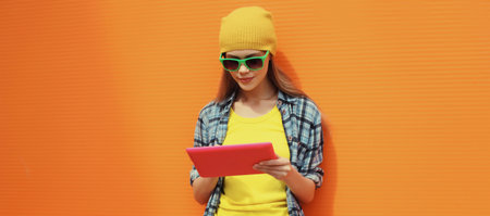 Portrait of modern stylish young woman using digital tablet computer, cool creative freelancer girl looking at device in colorful trendy clothes on city street on orange wall backgroundの写真素材