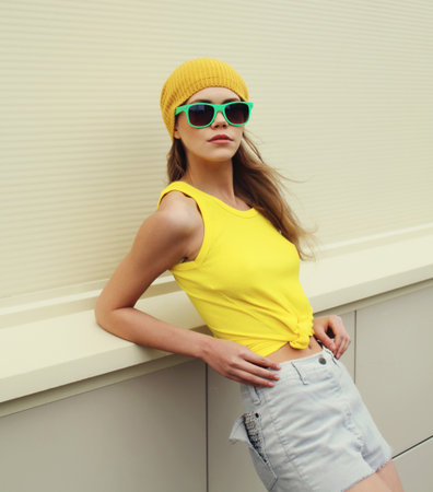Stylish young woman standing in colorful yellow hat, sunglasses, modern teenage girl posing on white wall background in the cityの写真素材