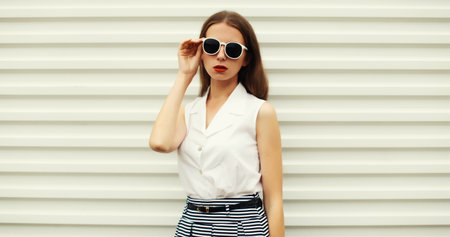 Fashion portrait of stylish young woman model in white glasses and shirt, modern girl posing on street wall backgroundの写真素材
