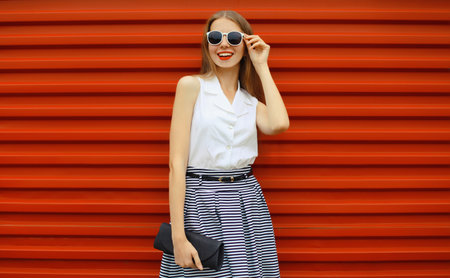 Beautiful happy stylish young woman in skirt with black handbag clutch, fashionable smiling girl standing on red street wall backgroundの写真素材