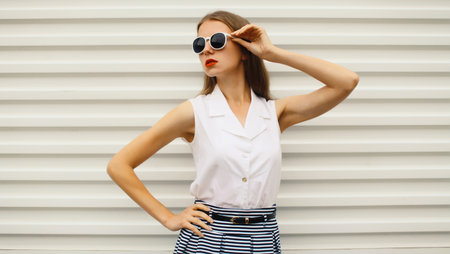Fashion portrait of stylish young woman model in white glasses and shirt, modern girl posing on street wall backgroundの写真素材