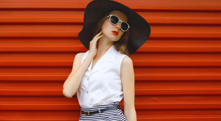 Beautiful stylish young woman in summer black straw hat, fashionable girl model standing on red street wall backgroundの写真素材