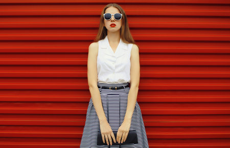 Beautiful stylish young woman in skirt with black handbag clutch, fashionable girl standing on red street wall backgroundの写真素材
