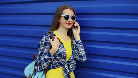 Happy young woman calling on mobile phone, modern girl talking on the smartphone in sunglasses looking away on vivid blue wall backgroundの写真素材