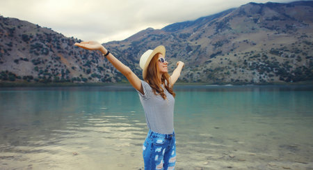 Summer vacation and tourism, happy smiling woman joyful tourist in straw hat raising her hands up on lake and mountains background. Greece, island Crete.の写真素材