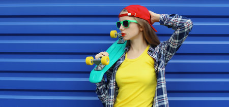 Stylish young woman posing with skateboard in colorful clothes, red baseball cap, cool modern teenage girl standing on vivid blue backgroundの写真素材