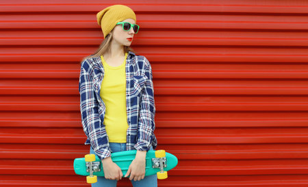 Stylish young woman posing with skateboard in colorful clothes, yellow hat, cool modern teenage girl standing on vivid red backgroundの写真素材