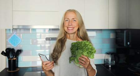 Cooking food, happy smiling woman holding phone with green lettuce vegetables using smartphone looks at a grocery list, a recipe for dish, or orders food for delivery standing in kitchen at homeの写真素材