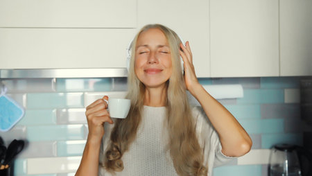 Portrait of happy smiling mature woman in headphones music with closed eyes enjoying hot tasty cup of coffee or tea standing in the kitchen in early morning at home, looking at cameraの写真素材