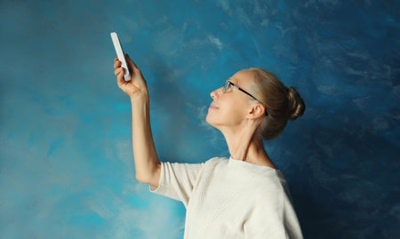 Happy smiling caucasian mature woman turns on the air conditioner using the remote control while standing in living room at homeの写真素材