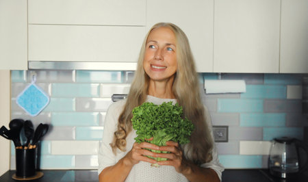 Cooking food, happy smiling mature woman with green lettuce vegetables looking away standing in the kitchen at home in the morningの写真素材