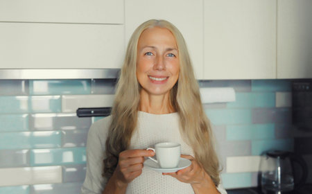Portrait of happy smiling mature woman enjoying hot tasty cup of coffee or tea standing in the kitchen in early morning at home, looking at cameraの写真素材