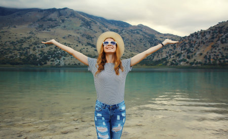 Summer vacation and tourism, happy smiling woman, joyful tourist in straw hat raising her hands up on lake and mountains background. Greece, island Creteの写真素材