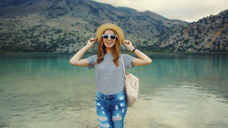 Summer vacation, happy joyful young woman tourist with backpack in straw hat standing on lake and mountains background. Greece, island Creteの写真素材