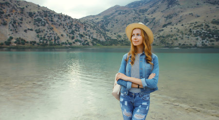 Summer vacation, young woman tourist looking away with backpack in straw hat standing on lake and mountains background. Greece, island Creteの写真素材