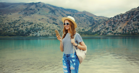 Summer vacation, happy young woman tourist using mobile phone wearing straw hat with backpack standing on lake and mountains background. Greece, island Creteの写真素材