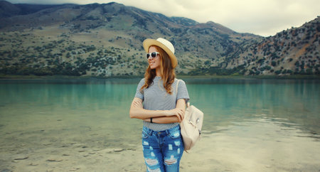 Summer vacation, happy young woman tourist with backpack in straw hat standing on lake and mountains background. Greece, island Creteの写真素材