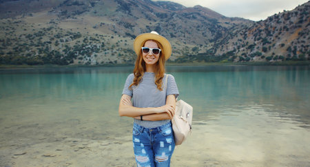 Summer vacation, happy young woman tourist with backpack in straw hat standing on lake and mountains background. Greece, island Creteの写真素材
