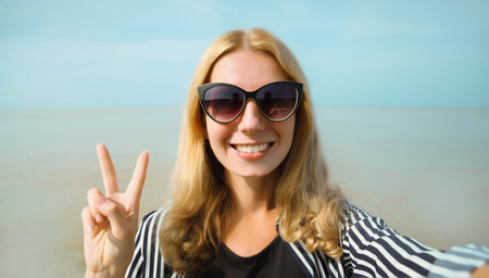 Summer portrait of happy cheerful young woman smiling taking selfie with smartphone on the beach, joyful girl laughing using phone on sea and blue sky backgroundの写真素材