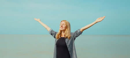 Summer vacation, happy inspired young woman raising her hands up on the beach, relaxed girl on sea coast with blue sky backgroundの写真素材