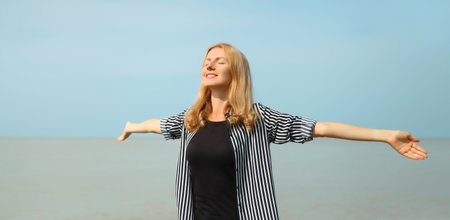 Summer vacation, happy inspired young woman raising her hands up on the beach, relaxed girl on sea coast with blue sky backgroundの写真素材