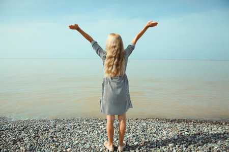 Summer vacation, happy inspired young woman raising her hands up on the beach, relaxed girl on sea coast with blue sky background, standing back viewの写真素材