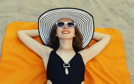 Summer vacation, beautiful happy relaxing young woman lying on sand on the beach wearing swimsuit, sunglasses with straw tourist hatの写真素材