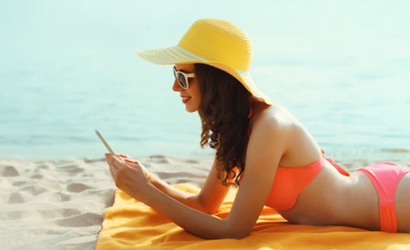 Summer vacation, happy young woman with phone, wear tourist hat, bikini, beautiful girl using smartphone lying on the sand on sea beach backgroundの写真素材