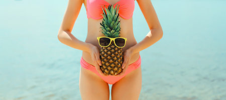 Summer vacation, close up beautiful young woman with pineapple in pink bikini swimsuit on the beach on sea coast backgroundの写真素材