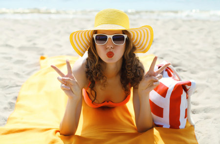 Summer vacation, happy cheerful young woman in bikini and straw tourist hat with sunglasses and bag lying on sand on sunny sea beach backgroundの写真素材
