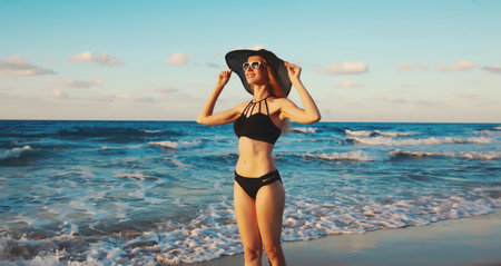 Summer vacation, beautiful slim young woman smiles in black swimsuit, tourist hat, sunglasses, happy smiling girl standing on beach, sea coast background with waves at sunset, tropical islandの写真素材