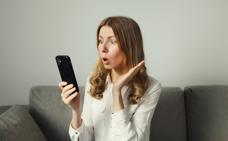 Emotional surprised young woman looking at phone, girl with smartphone sitting on the couch in...