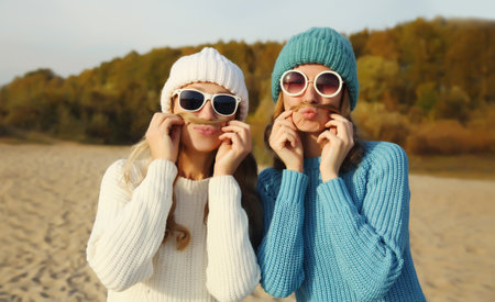 Portrait of happy cheerful two women friends having fun showing mustache in sunglasses, winter colorful hat, glasses, funny girlfriends together outdoorsの写真素材