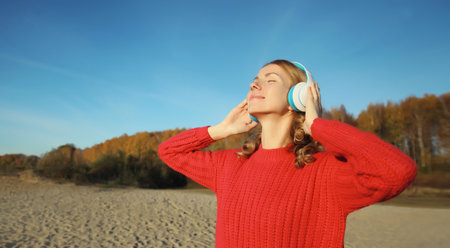Happy relaxed young woman enjoying listening to music in headphones inspired outdoors on autumn coast by sea against trees, life momentの写真素材