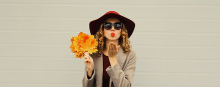 Autumn portrait of beautiful young woman with yellow maple leaves blowing a kiss in hat, red lipstick, posing on gray wall backgroundの写真素材
