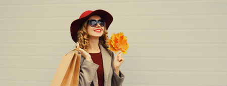 Autumn portrait of beautiful happy young woman with shopping bag, stylish girl holding yellow maple leaves in round hat, coat on gray studio backgroundの写真素材