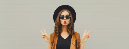 Portrait of beautiful young woman blowing a kiss, stylish girl in black round hat posing on gray backgroundの写真素材