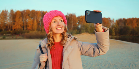Travel, adventure, happy traveler young woman taking selfie with phone on the beach at sunset, girl using smartphone standing on sea or river coast with forest, trees, landscape, nature backgroundの写真素材