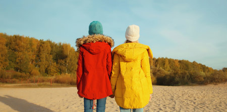 Back view of full length two young modern women friends standing together on sunny beach, stylish...