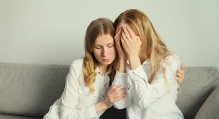 Upset offended caucasian two women, mature mother or sister and adult daughter crying together sitting on couch at homeの写真素材
