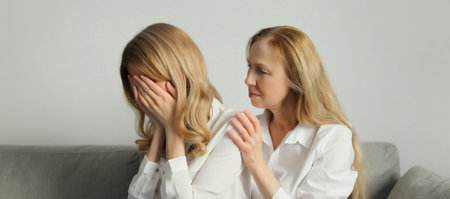 Upset offended caucasian two women, mature mother or sister and adult daughter crying together sitting on couch at homeの写真素材