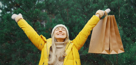 Happy smiling young woman with shopping bag in winter against Christmas tree, joyful girl in yellow jacket, hat standing in snowy forestの写真素材