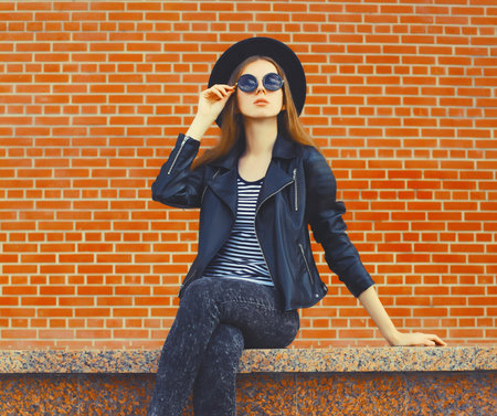Portrait of stylish young woman model wearing black round hat, leather jacket in rock style on city street over brick wall backgroundの写真素材