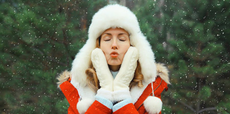Happy smiling young woman having fun enjoys snow, warm weather in winter park against Christmas tree, joyful girl in white hat standing in forest with snowflakes, festive backgroundの写真素材