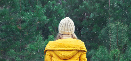 Back view, winter day, happy young woman enjoys snow, warm weather, joyful girl in hat, yellow jacket in snowy green forest with snowflakesの写真素材