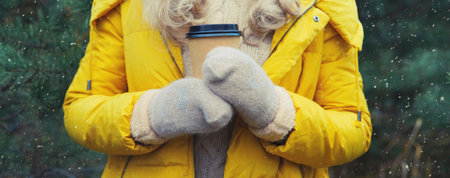 Close up female hand holding cup of hot coffee drink against Christmas trees in winter forest, wearing yellow jacket, mittensの写真素材