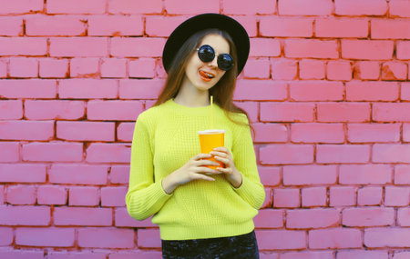 portrait of happy stylish young woman in black hat, yellow sweater, trendy modern girl with juice drink cup posing on colorful pink wall urban backgroundの写真素材