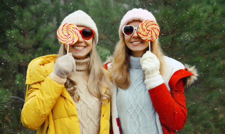 Portrait of happy smiling women friends having fun, celebration, joyful girlfriends together with sweet lollipop on Christmas tree background in winter forest, wearing hat, colorful jacket outdoorsの写真素材