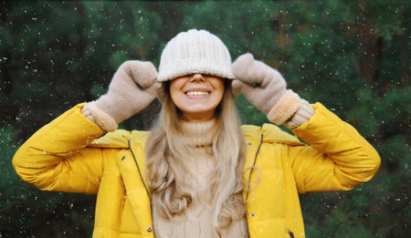 Happy cheerful young woman having fun enjoys snow, warm weather in winter park against Christmas tree, joyful girl in hat standing in forest with snowflakes, festive backgroundの写真素材